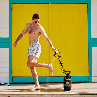 A smiling man in sunglasses and striped swim shorts uses a black Beach Soul portable shower to rinse his feet on a wooden deck in front of a vibrant yellow and turquoise beach hut