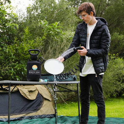 Photograph of a young person wearing clear glasses and a black puffer jacket washing a white plate with a black Beach Soul portable shower on a camping table next to a green camp stove and a khaki tent in a lush green outdoor setting