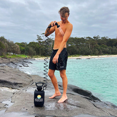 Photography of a shirtless young man in black Calvin Klein shorts rinsing off with a black Beach Soul portable shower on a rocky coastline next to vibrant turquoise water and a sandy beach lined with lush green trees under a cloudy sky