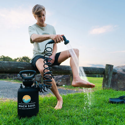 A young man in a light green t-shirt and black shorts sits on a rustic wooden fence washing his bare feet with a black Beach Soul portable shower featuring an orange sun and blue wave logo on vibrant green grass under a clear blue sky