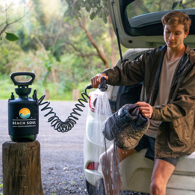 A young man sits on the tailgate of a white car, washing his muddy hiking boots with a black Beach Soul portable shower featuring an orange wave logo and coiled hose, set against a lush green outdoor background