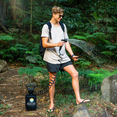 Photography of a young man wearing sunglasses and a backpack demonstrating a black Beach Soul portable shower with a coiled hose spraying water onto lush green ferns in a dense forest