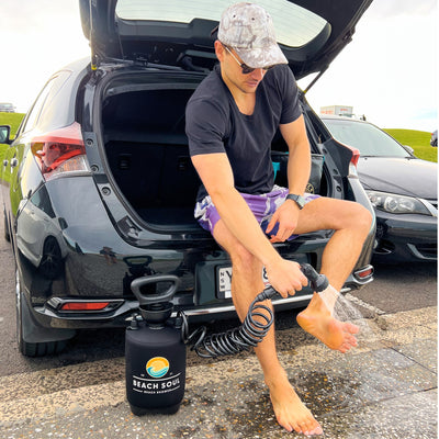 A man in a camouflage hat and purple patterned swim shorts sits on a black car's open trunk, washing his bare feet with a Beach Soul portable shower in New South Wales Australia
