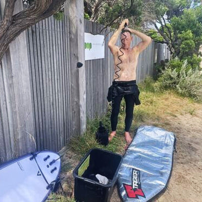 A shirtless man in black wetsuit bottoms showers with a coiled hose near a weathered wooden fence, with a pale blue surfboard and a silver TRAGER board bag resting on the sandy ground