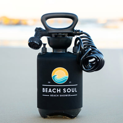 Photography of a black portable Beach Soul beach shower with a distinctive yellow sun and blue wave logo, a coiled hose, and an adjustable nozzle, positioned on a sandy shore
