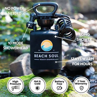 A black Beach Soul portable shower with a blue and orange wave logo, a coiled hose, and a 10-mode showerhead, displayed on green mossy rocks in a blurred outdoor setting