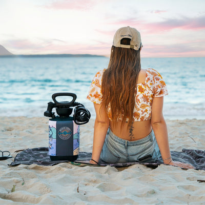 Photography by unknown artist of a person on a sandy beach at sunset, viewing the turquoise ocean and a faint mountain, wearing a white floral crop top and beige Capetown cap, with a colorful Beach Soul portable shower beside them