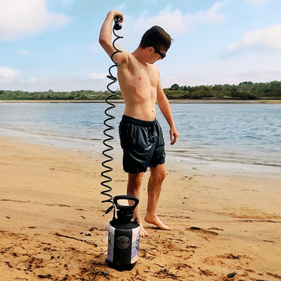 A young man in black swim trunks and sunglasses showers with a portable unit featuring a black coiled hose and colorful sleeve on a sandy beach with calm water and green trees in the background