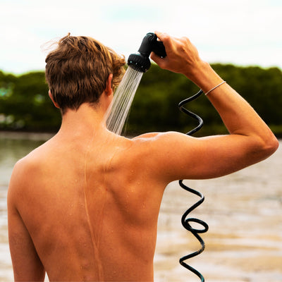 A person showering their head and bare back outdoors near a body of water with green trees, holding a black hand-held shower sprayer, with water visibly streaming down their tanned skin and a silver bracelet on their right wrist