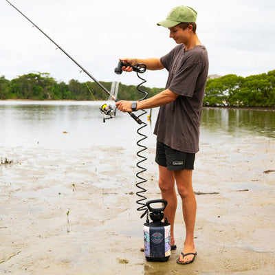 A young man in a green corduroy cap cleans a fishing rod with a yellow line using a portable shower featuring a coiled black hose and a colorful insulated cover on a muddy shoreline with green trees in the background
