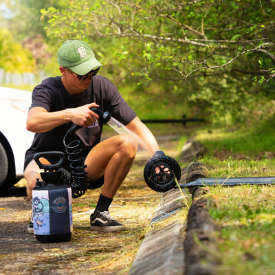 A person in a green corduroy cap embroidered with a face and black Vans sneakers uses a portable Beach Soul shower, adorned with a colorful surfer and fish graphic, to rinse a dirty black beach cart wheel on an outdoor path