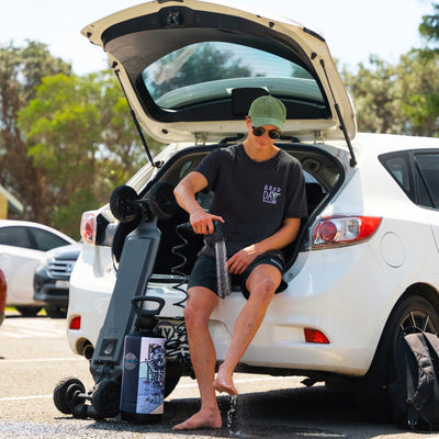 Photograph of a young man wearing a green cap and a "Good Day To Explore" t-shirt cleaning his bare feet with a portable shower from the open trunk of a white car in a sunny parking lot next to an electric skateboard and a water bottle featuring a skeleton graphic