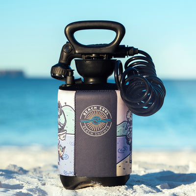 Product photography of a black portable Beach Soul Beach Shower on white sand, featuring a colorful neoprene sleeve with beach cartoons and a sun-wave logo, against a turquoise ocean and clear blue sky
