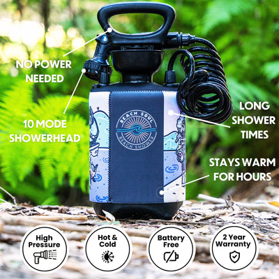 A black Beach Soul portable pump shower with a vibrant neoprene sleeve featuring a sun and wave logo and playful cartoon beach graphics stands on natural ground amidst green foliage, highlighting its coiled hose and 10-mode showerhead as a battery-free solution