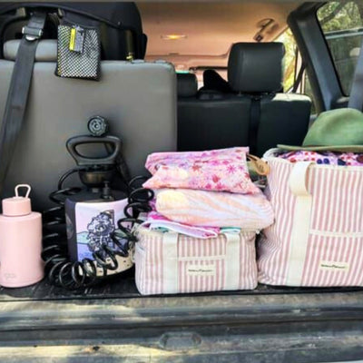 A car trunk packed for a trip with pink and white striped tote bags, a pastel pink water bottle, a portable pressurized shower, and a green floppy hat