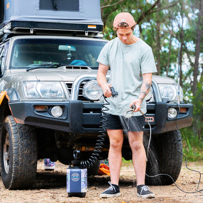 A man in a peach cap and light green Assembly Label t-shirt showers beside a tan Nissan Patrol SUV with a grey rooftop tent and ARB bull bar, actively using a portable Beach Soul Beach Shower unit in a natural forest setting