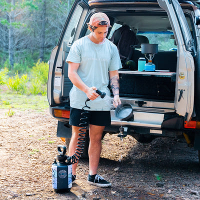 Photography of a young man wearing an ASSEMBLY LABEL t-shirt and coral cap, rinsing a metal pan with a portable shower next to an open beige SUV, revealing a blue camping stove and thermos inside, while a BEACH SOFT portable shower pump rests on the gravel ground