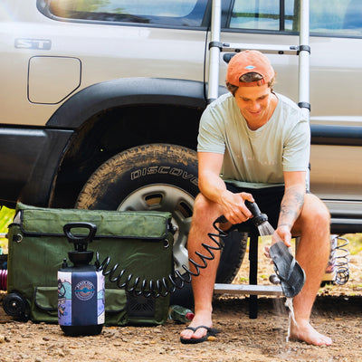 A man wearing an orange cap and light green t-shirt sits by a beige Nissan Patrol ST 4x4 with a dirty Discover tire, using a portable Beach Soul Shower bottle with a blue and purple graphic to rinse his black flip-flops on a dusty ground next to an olive green collapsible wagon
