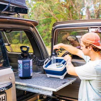 Photography of a young man in an orange cap washing items with a portable camping shower featuring a colorful graphic sleeve and a blue collapsible basin, set up on a pull-out table from his SUV at a wooded campsite with a roof-top tent