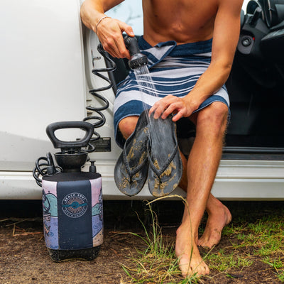 A person wearing a blue and white striped towel sitting by a white car, using a Beach Soul portable shower with a colorful illustrated sleeve to rinse sand-covered black flip-flops, with their bare muddy feet visible on the dirt ground