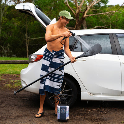 A shirtless young man wearing a green cap and a blue and white striped towel rinses a black paddleboard paddle with a coiled hose from a branded Beach Soul portable shower beside a white car with its trunk open on muddy ground
