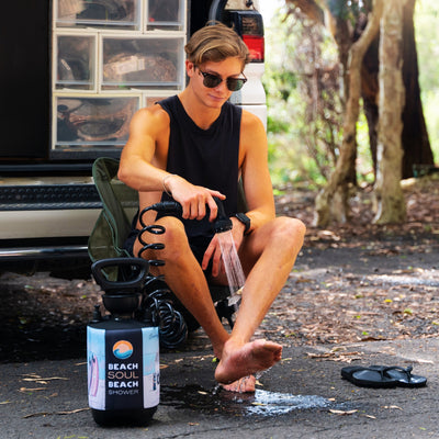 A young man in sunglasses washes his bare feet with a portable Beach Soul Beach Shower unit while seated in a green camping chair beside an open white camper van revealing clear storage containers at a shaded campsite