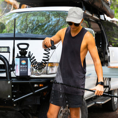 Digital photograph of a man in a Happy Days cap and sunglasses operating a black Beach Soul portable shower with a blue and orange label to spray water on a long black rod beside a white Mitsubishi Pajero off-road vehicle equipped with a roof rack and bull bar outdoors