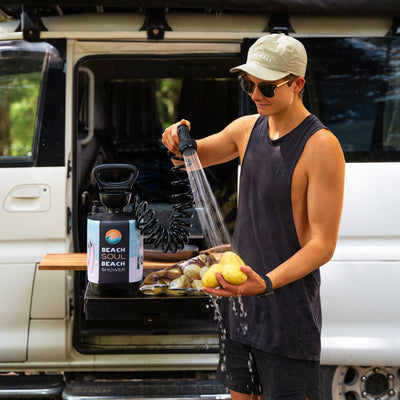 A person wearing a beige "Happy Days" cap and sunglasses washes yellow potatoes with a black coiled hose from a "Beach Soul Beach Shower" unit next to a white camper van with a wooden side table