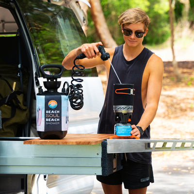 Photography of a young man in sunglasses and a black tank top pouring water from a "Beach Soul Beach Shower" into a portable camping stove with bright orange handles, set on a wooden pull-out counter from a camper van outdoors