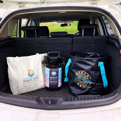 Photography of Beach Soul brand beach day essentials including a white tote bag a black portable shower with surfboard graphics and a black collapsible bucket with bright blue straps all packed in a car trunk