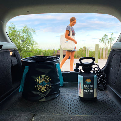 A woman carrying a white Beach Soul tote bag walks on a sandy path past wooden fence posts and a wooden bench as seen from a car trunk holding a black collapsible bucket with a gold sun logo and a portable shower with a colorful surfboard sleeve