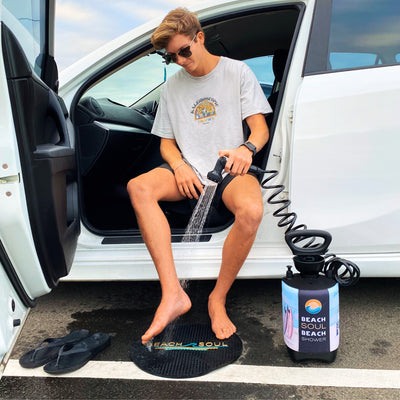 A young man in sunglasses and a Kaleidoscopic Tropics t-shirt sits in the open door of a white car washing his sandy feet with a portable Beach Soul Beach Shower onto a black Beach Soul mat in a parking lot