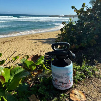 A light blue portable shower bottle with a cartoon vintage van and surfers rests on vibrant green coastal foliage with a dry brown leaf nearby, set against a golden sandy beach with crashing blue ocean waves and distant rocky headlands under a clear sky