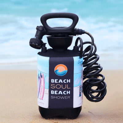 Photography, a black portable Beach Soul Beach Shower with a colorful neoprene sleeve by Benjamins featuring an orange sun and blue wave logo, pink surfboards, and "BEACH SOUL BEACH SHOWER" text stands on a sandy beach against a turquoise ocean with a visible black coiled hose and adjustable spray nozzle