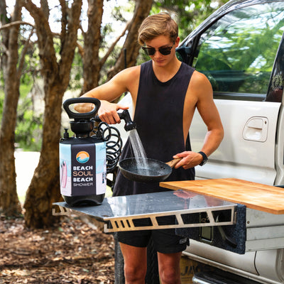 A young man in a black tank top and sunglasses rinses a speckled frying pan with a blue Beach Soul Beach Shower featuring pink surfboards, set up next to a white overland vehicle with a pull-out kitchen table in a leafy outdoor environment