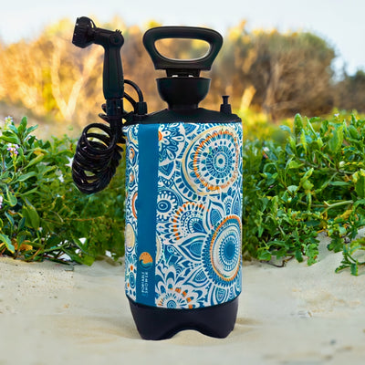 Photograph of a portable shower with a vibrant blue and orange mandala pattern standing on light sand amidst green beach vegetation, featuring a black nozzle, coiled hose, and a blue vertical band that reads PORTABLE SHOWER