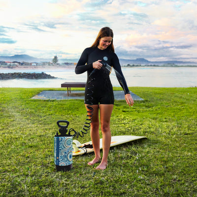 A young woman in a black shorty wetsuit rinses off with a portable blue mandala-patterned shower, her surfboard resting on the grass beside her, at a coastal park overlooking a rock wall, waves, and distant town against a cloudy, pastel sky