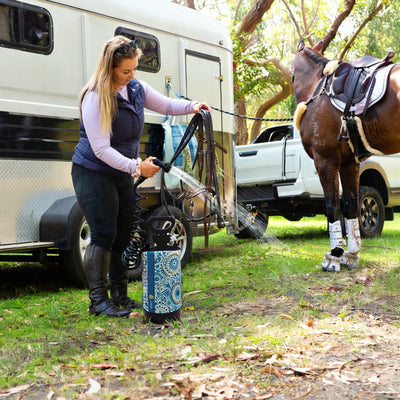 A woman in a purple long-sleeve and navy vest hoses down brown leather horse tack using a portable washer with a blue and white mandala pattern, next to a brown horse wearing a western saddle and white leg boots by a white trailer and pickup truck in a grassy outdoor setting