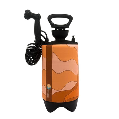 Product photography of a cylindrical portable shower unit with a wavy orange and brown desert pattern, a black pump handle, a coiled black hose with a shower head, and a side label featuring the Portable Shower logo