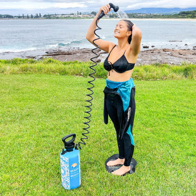 A smiling woman in a black bikini and wetsuit rinses her hair with a blue portable Beach Soul Shower, standing barefoot on a black mat on green grass overlooking a rocky coastline and distant sandy beach