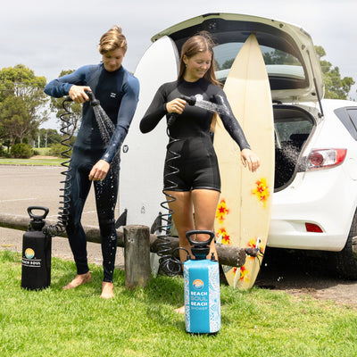 Two surfers in wetsuits use portable Beach Soul showers to rinse off next to a white car with two surfboards, one adorned with red and yellow star decals, in a grassy beach parking lot
