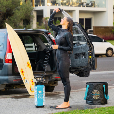 Photograph of a woman in a black wetsuit smiling while showering with a turquoise Beach Soul portable pressure shower on a residential street, a yellow surfboard with red star designs leaning against a black SUV and a black Beach Soul bucket next to her