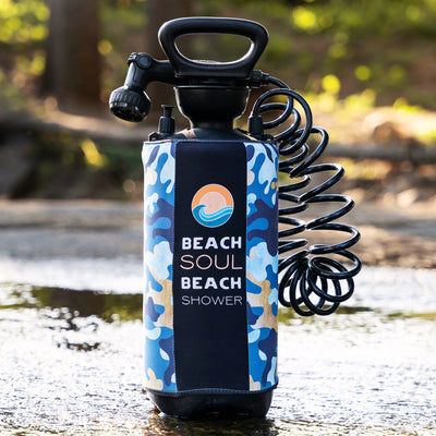A portable black sprayer, the Beach Soul Beach Shower, with a blue white and tan camouflage sleeve featuring an orange sun and blue wave logo with product name text, stands in shallow glistening water