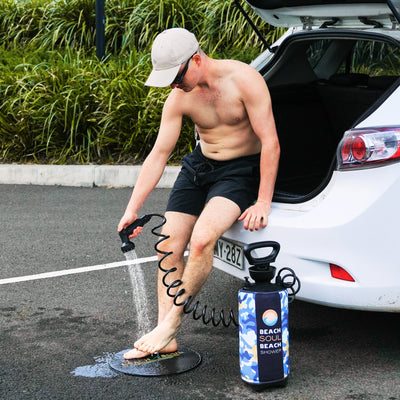A shirtless man in a beige cap and black shorts sits on the open trunk of a white car washing his feet with a portable Beach Soul Beach Shower with a blue camouflage pattern onto a black textured mat in a parking lot