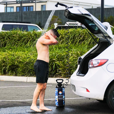Photography of a shirtless man in black shorts with water spraying over his head from a portable blue camouflage Beach Soul Beach Shower connected to an open white car trunk in an outdoor parking area