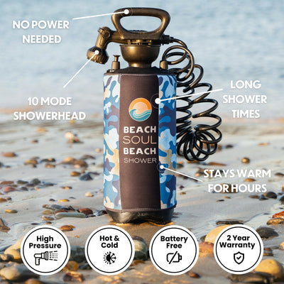 A Beach Soul portable shower in a blue camouflage neoprene sleeve with its brand logo, black pump handle, and coiled hose stands on a sandy pebbly beach, featuring a 10-mode showerhead and battery-free design