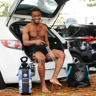 A smiling shirtless man uses a portable Beach Soul shower to rinse his legs while sitting on the open trunk of a white car, with a blue camouflage shower bottle and a black Beach Soul bucket with turquoise handles on the ground covered in brown leaves