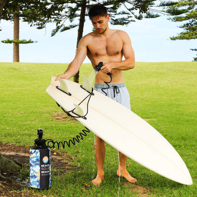 A shirtless toned man in light blue board shorts uses a blue camouflage portable Beach Soul Beach Shower to rinse a white surfboard with three fins in a bright grassy park