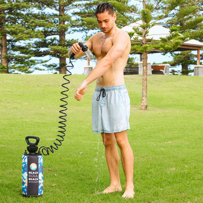 A shirtless man in light blue swim shorts showers his arm with water spraying from a black nozzle connected by a spiral hose to a blue camouflage Beach Soul portable shower in a grassy park featuring tall pine trees and white gazebos