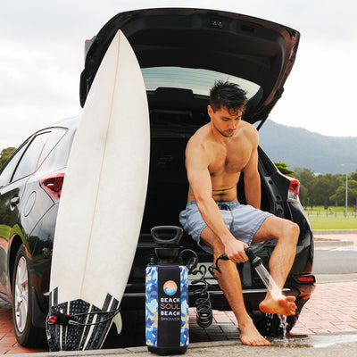 A man in grey shorts showers his sandy feet with a blue camouflage Beach Soul Beach Shower next to an open black SUV and a white surfboard with black grip pads against a backdrop of green parkland and distant mountains
